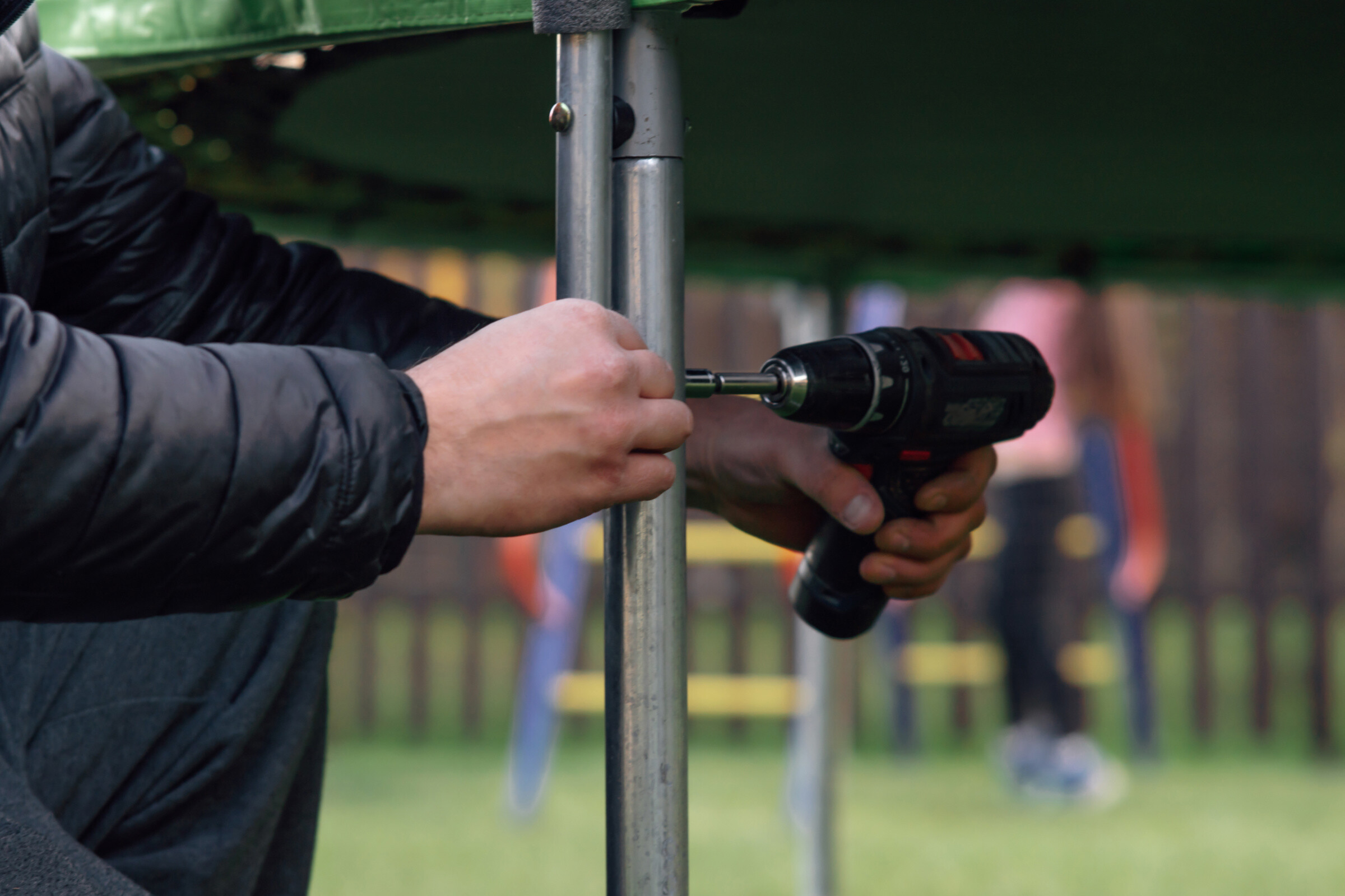 A man finishing up the assembly of a trampoline by drilling a screw into the metal trampoline leg.