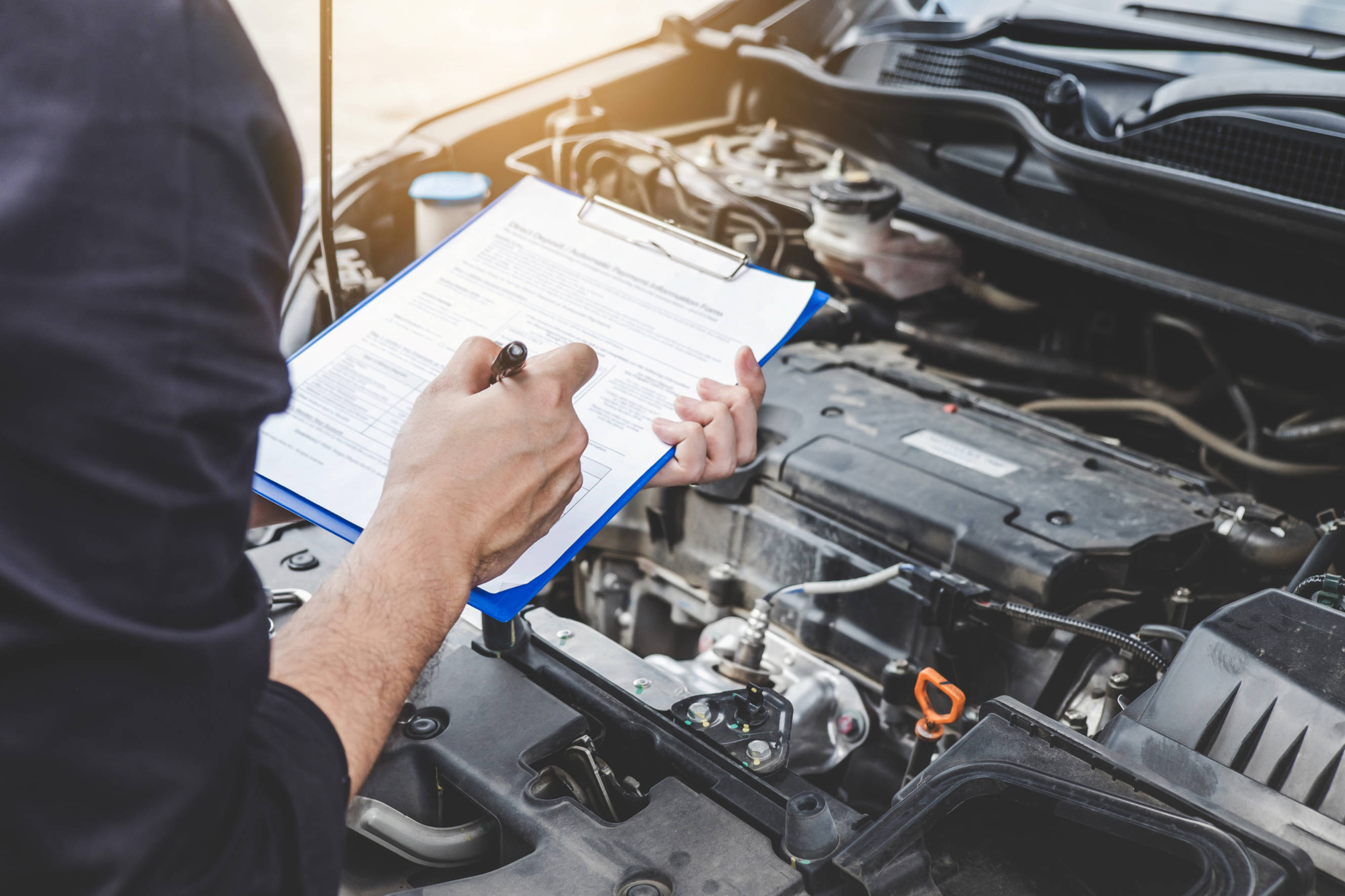 Mechanic performing car diagnostics with a checklist in hand while inspecting the engine compartment.