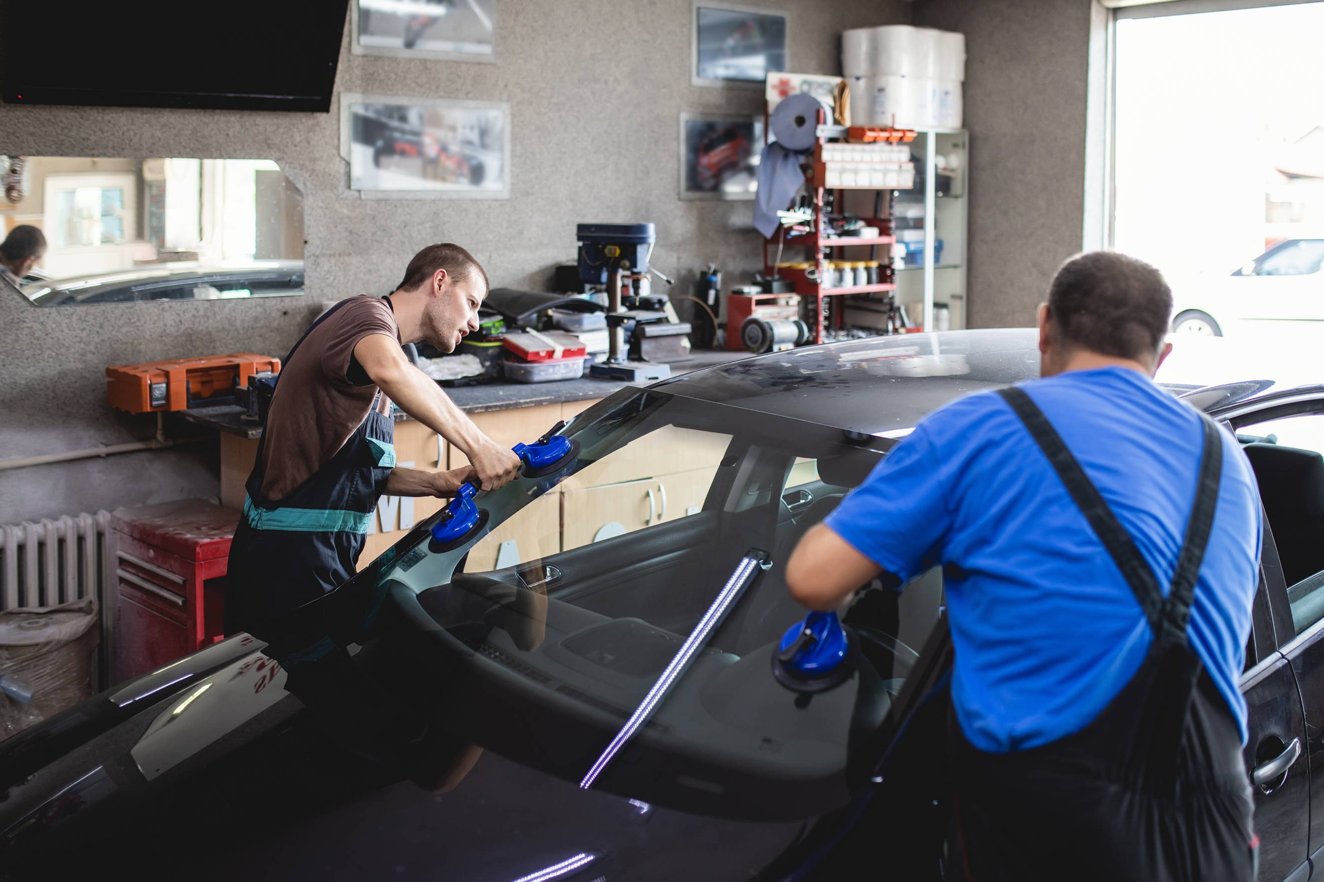 Two mechanics performing car window replacement using specialized suction tools to install a new windshield in a workshop.