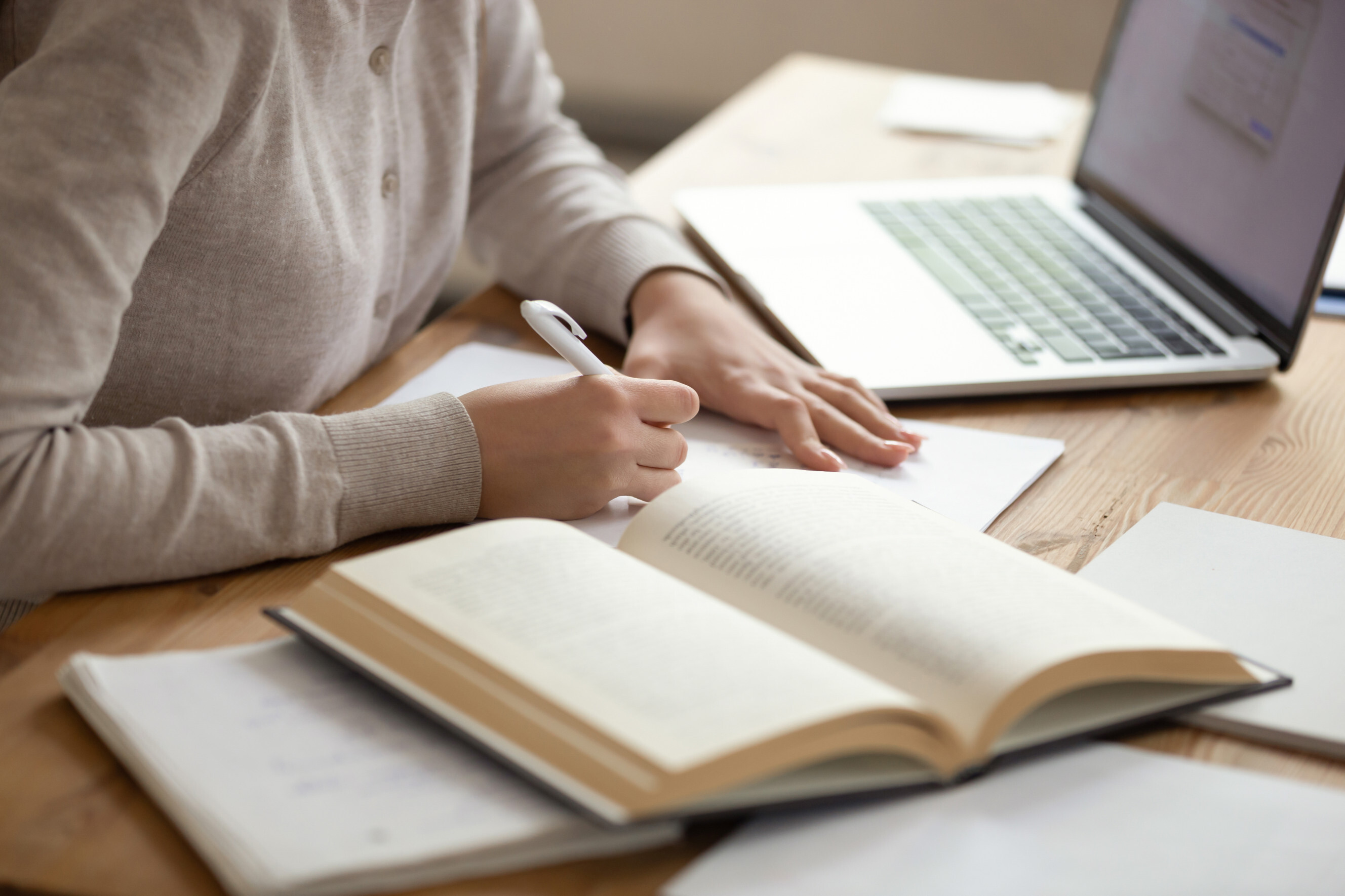 Person writing notes in a notebook with an open book and laptop on the desk, representing academic writing services.