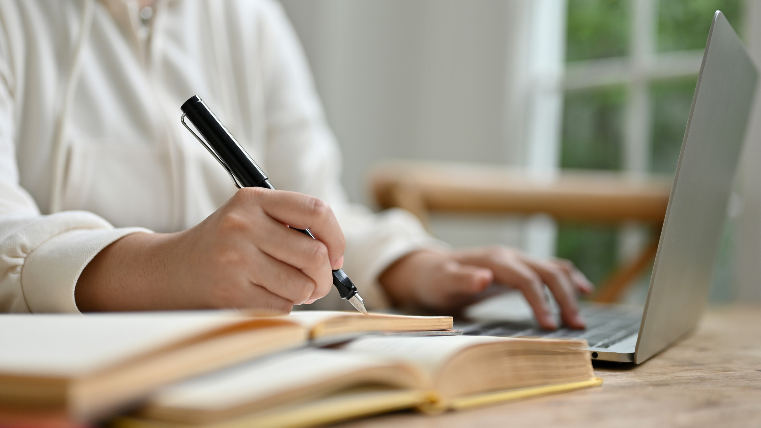 Person writing in a notebook with an open book and typing on a laptop, symbolizing article writing services.