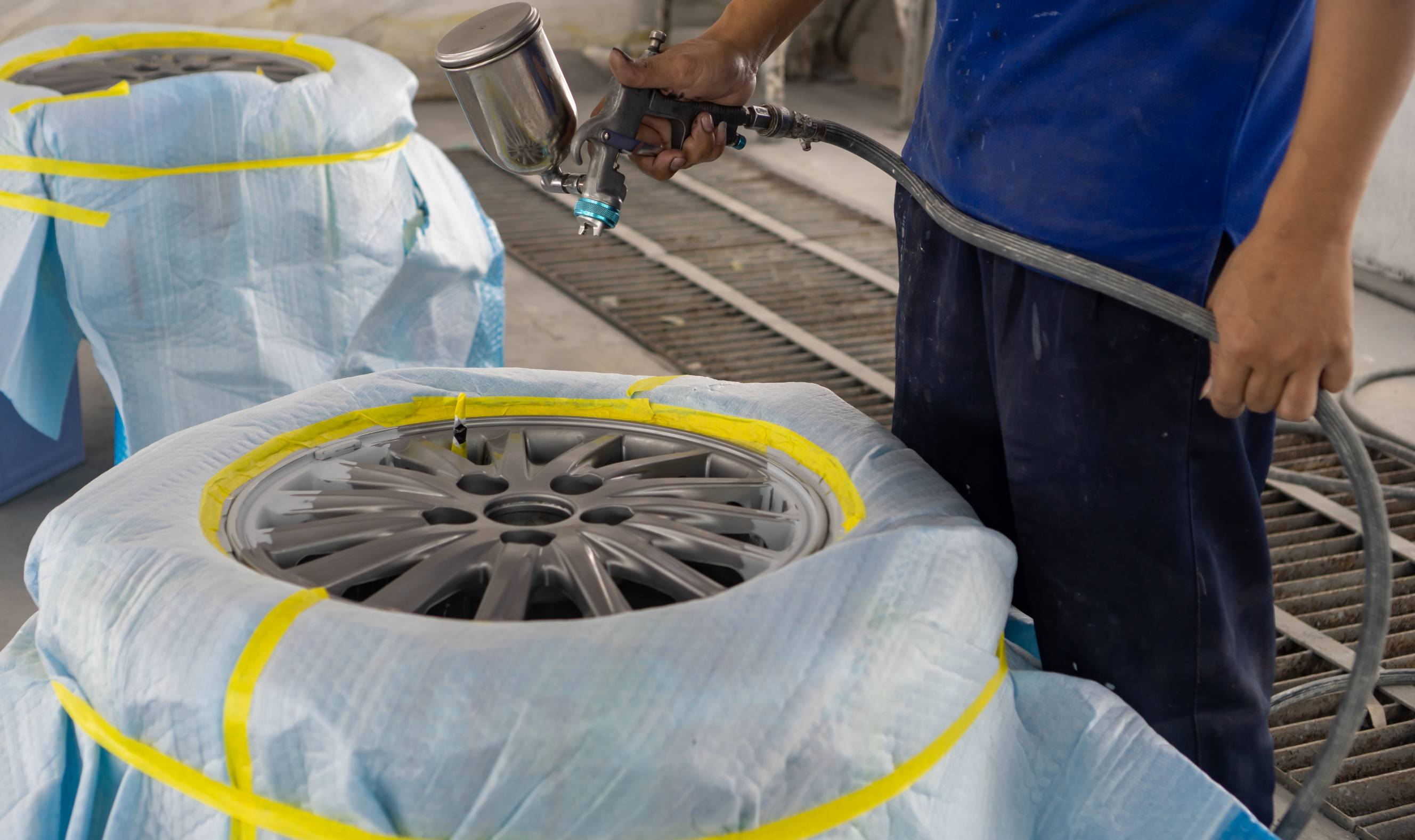 A mechanic performing wheel refurbishment by spray painting a car rim in a workshop.