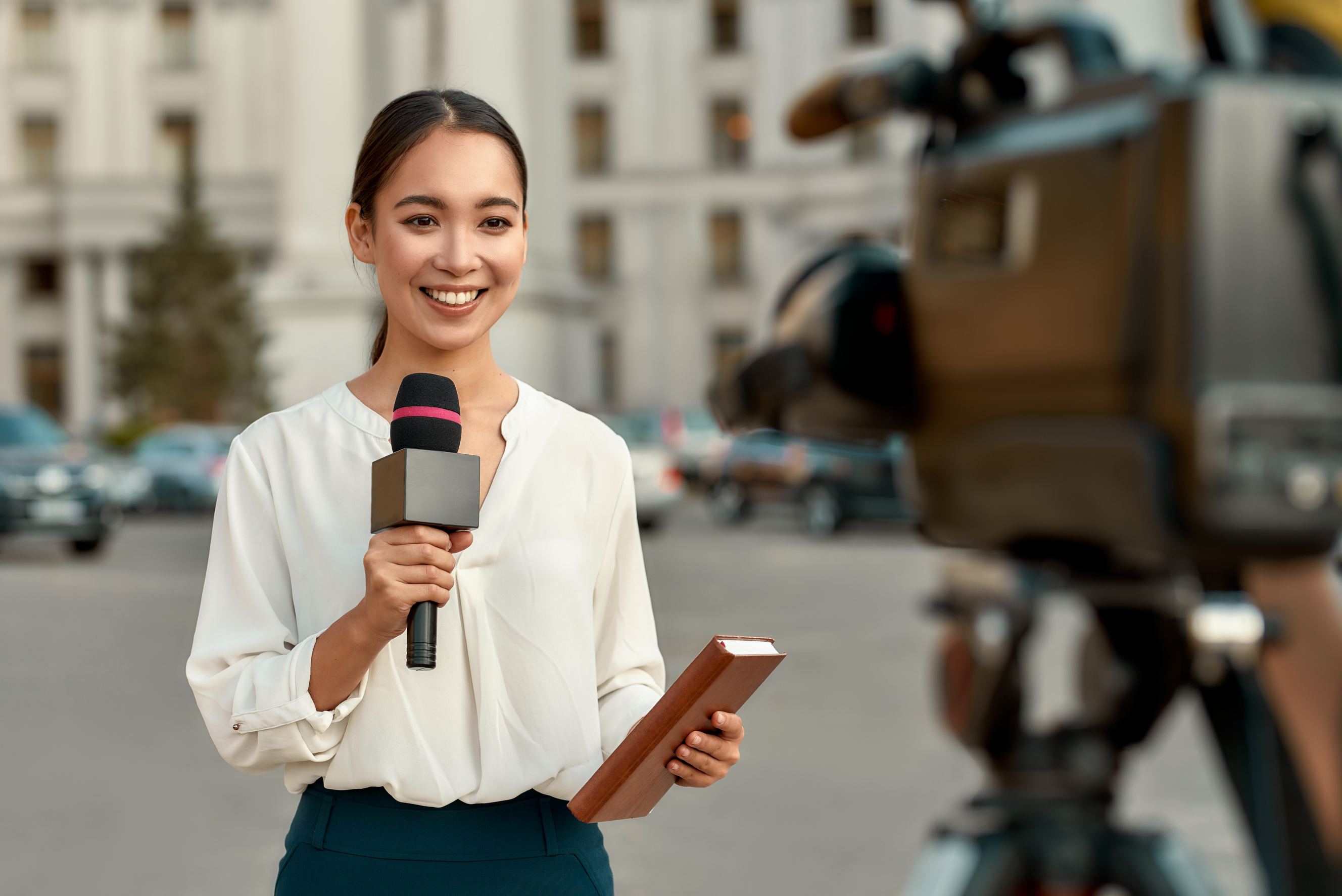 Young Asian lady journalist standing on the street with a microphone in hand and smiling at the camera.