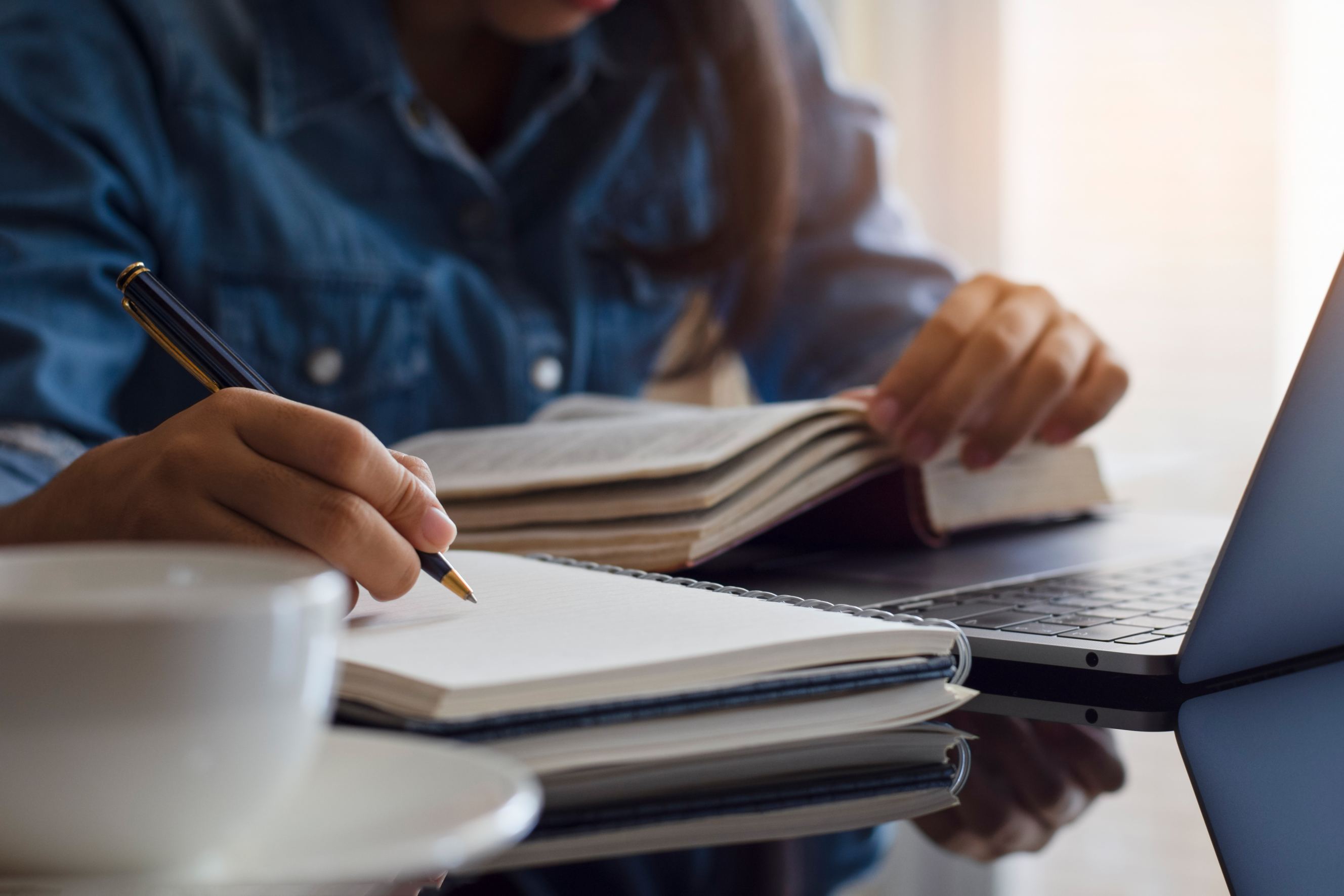 A casual young female selection criteria writer with a notebook while holding a book beside her laptop.