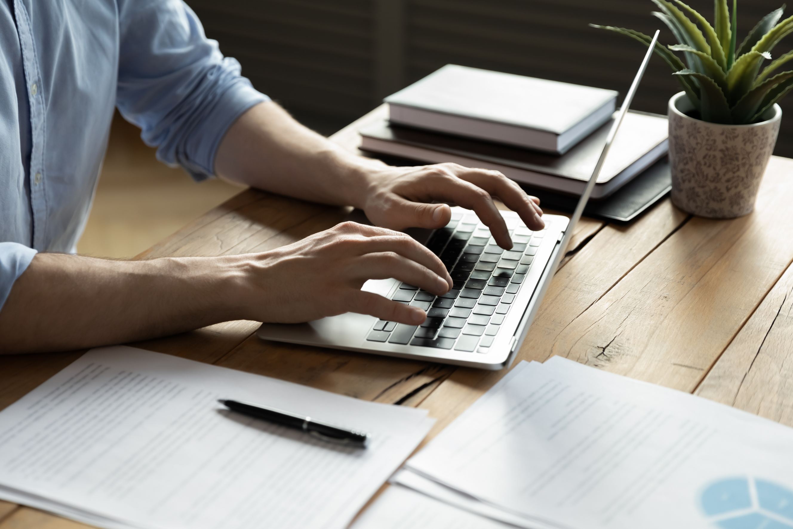 A close-up speech writer typing on a laptop, sitting at a wooden desk with documents,