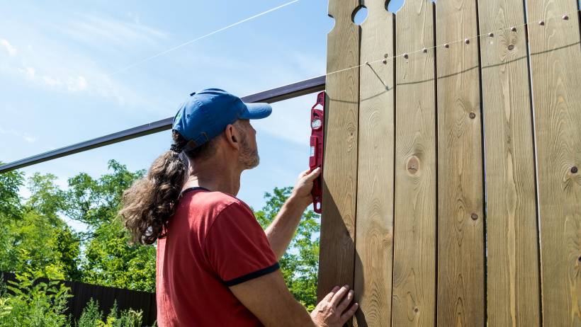 A person, wearing a blue cap and red shirt, is using a power tool to attach wooden boards to a fence in an outdoor setting. - fencing cost