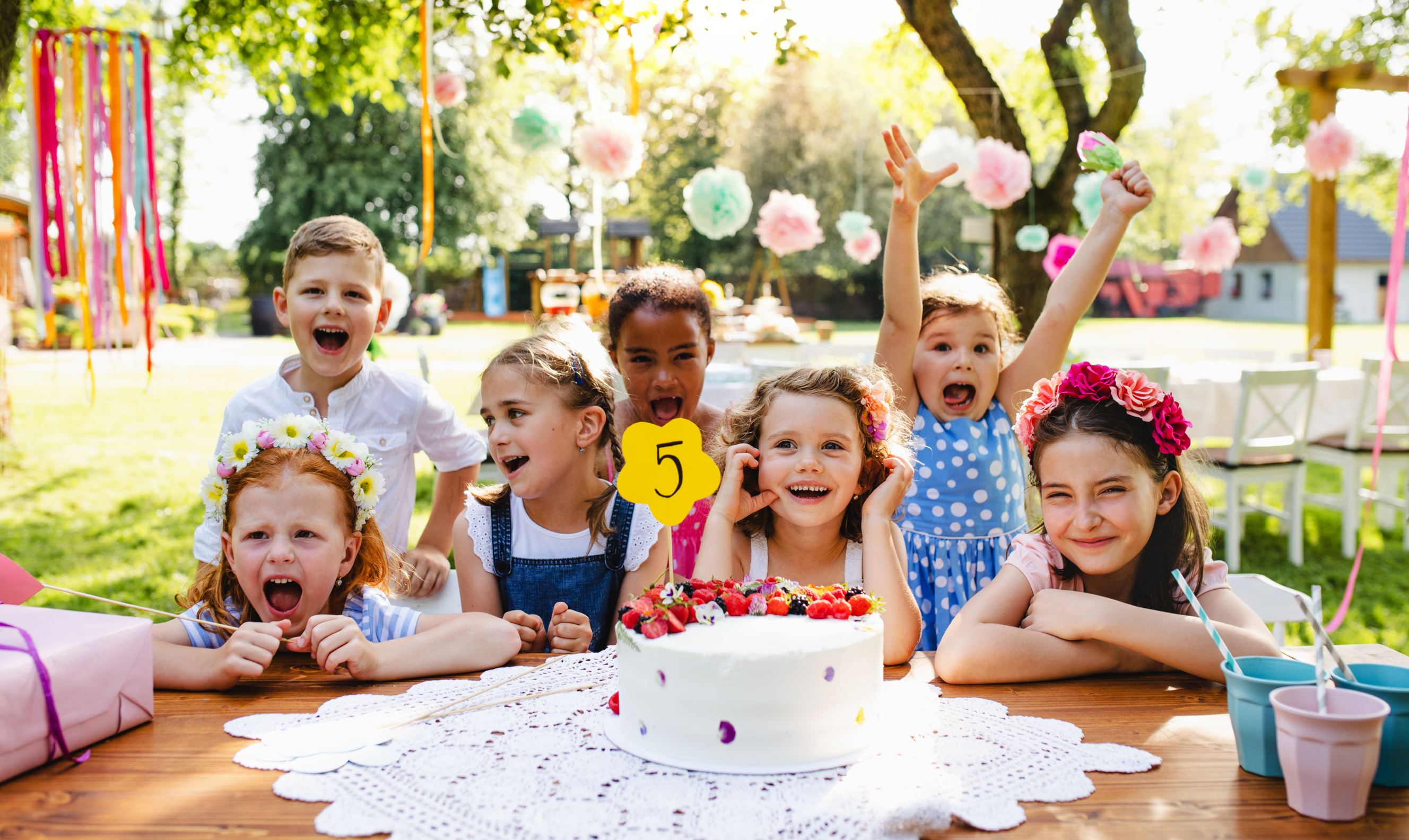 A portrait of children with cake standing around the table at a birthday party in the garden in summer taken by birthday photographer.