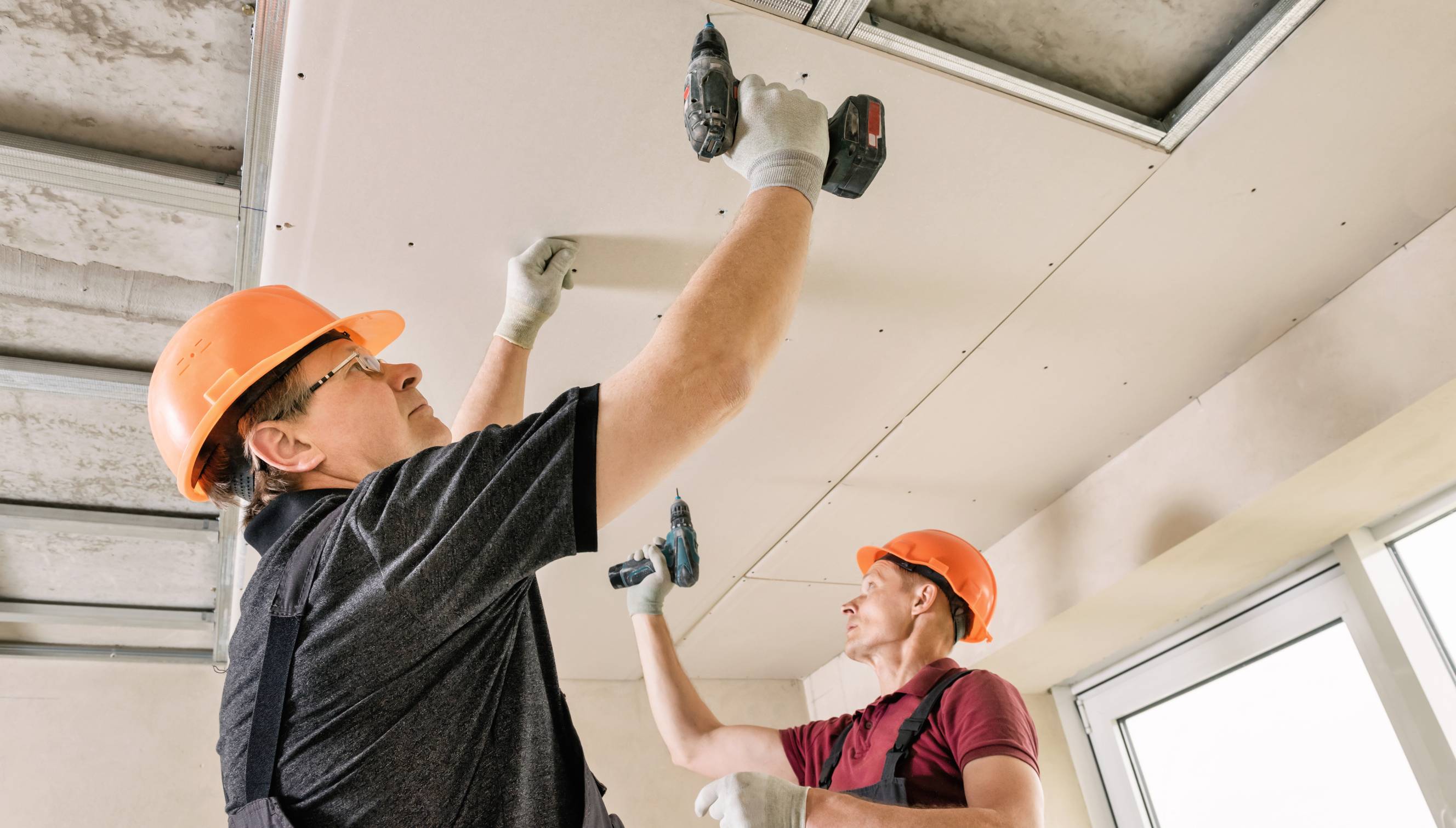 Handymen installing sheetrock drywall panels on a ceiling using power tools. 