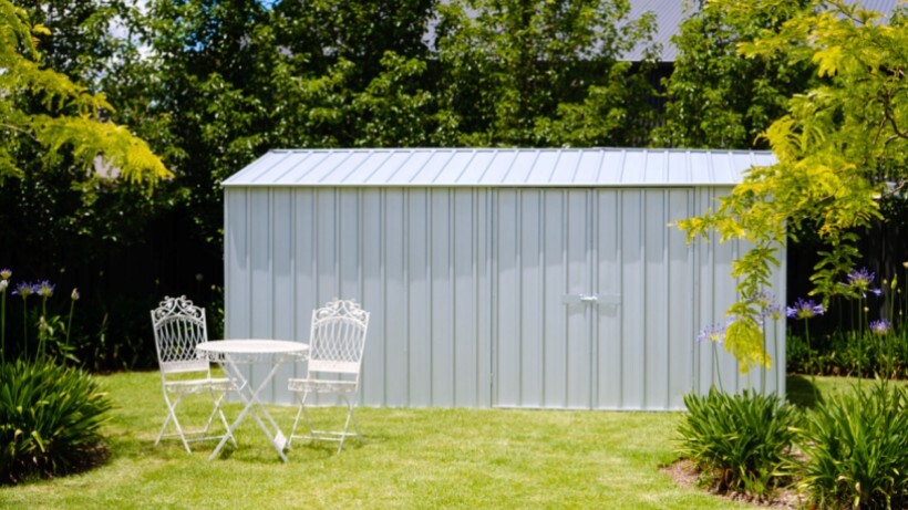 A neatly installed gray garden shed surrounded by lush greenery, with a white outdoor table and chairs set in front for added charm.