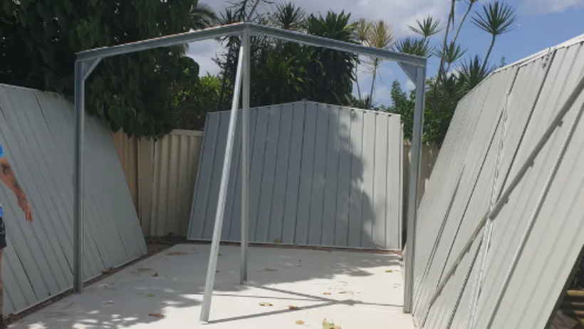Partially constructed shed with metal framing and detached panels, surrounded by greenery in a backyard