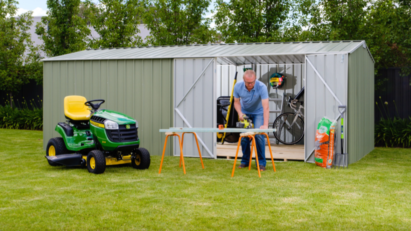 A green workshop shed by Easyshed with open doors, showing tools and equipment inside, and a man working on a project in front with a ride-on mower nearby