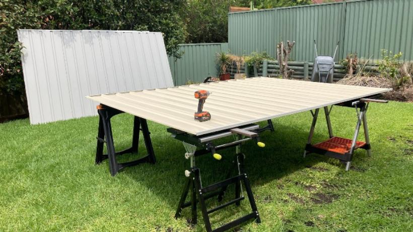 Shed installation in progress with metal panels laid on sawhorses, a drill placed on top, and a green backyard with fencing in the background