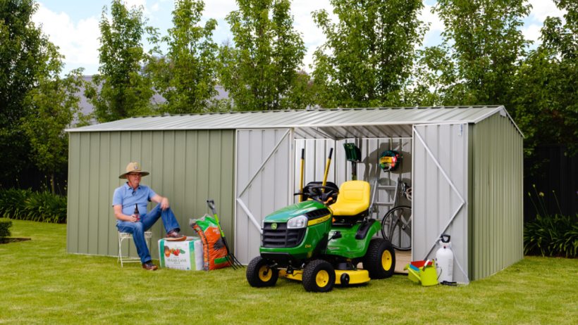 Timber frame shed vs steel shed -  A spacious green steel shed by EasyShed, open to showcase gardening equipment, a lawnmower, and tools, with a man sitting nearby in a lush backyard setting