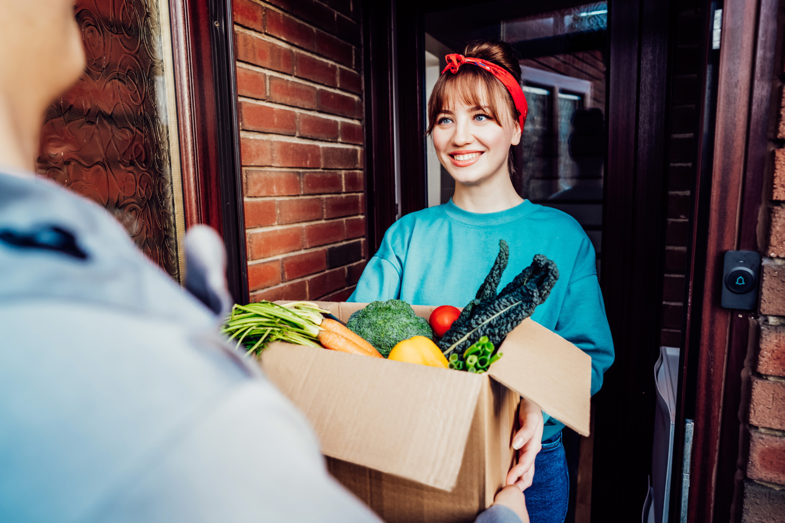 Smiling delivery person handing a box of fresh vegetables to a customer at their doorstep, highlighting reliable vegetable deliveries near you.