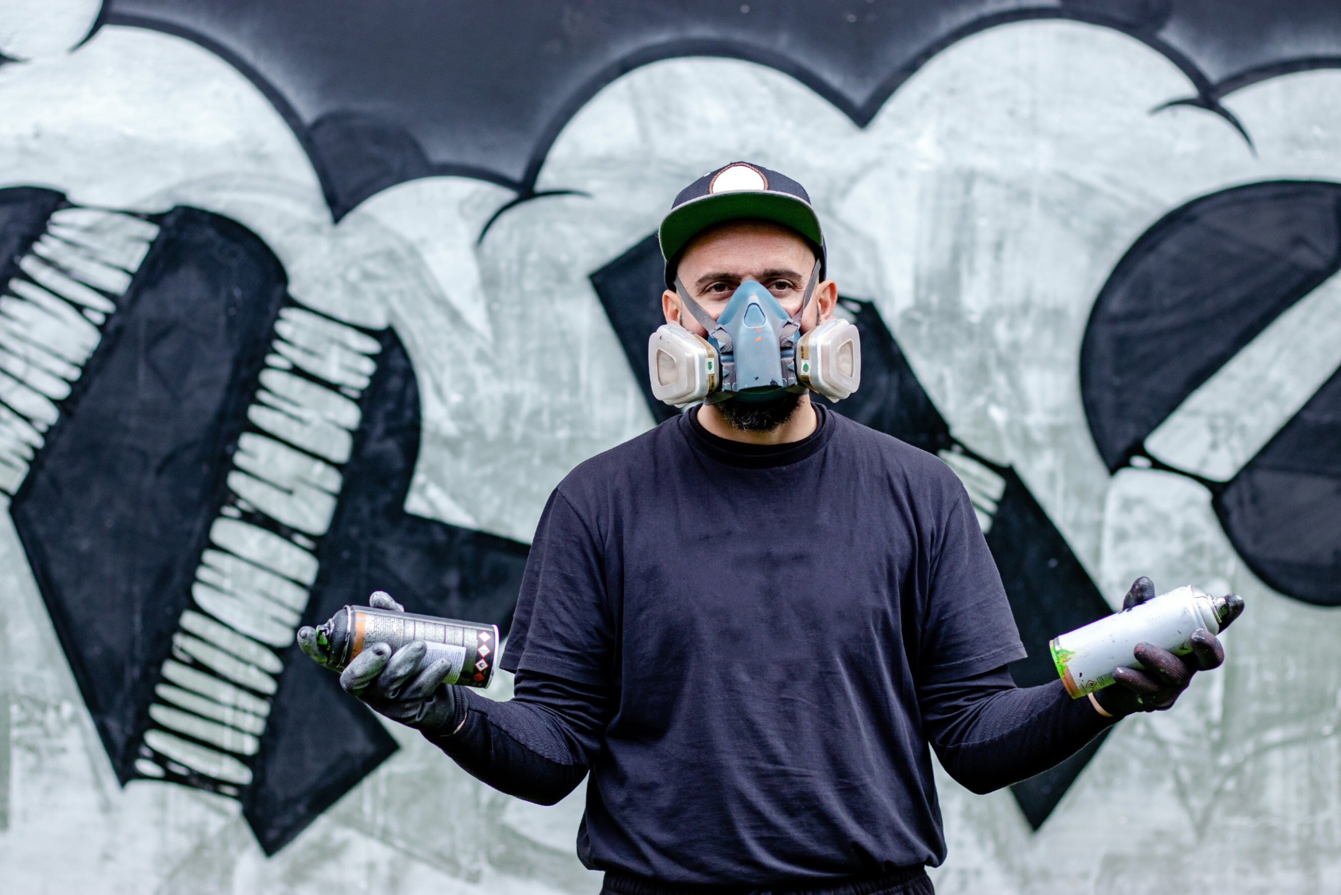 Graffiti artist wearing a respirator mask holding spray paint cans, standing in front of a creative mural, showcasing professional graffiti artists near you.