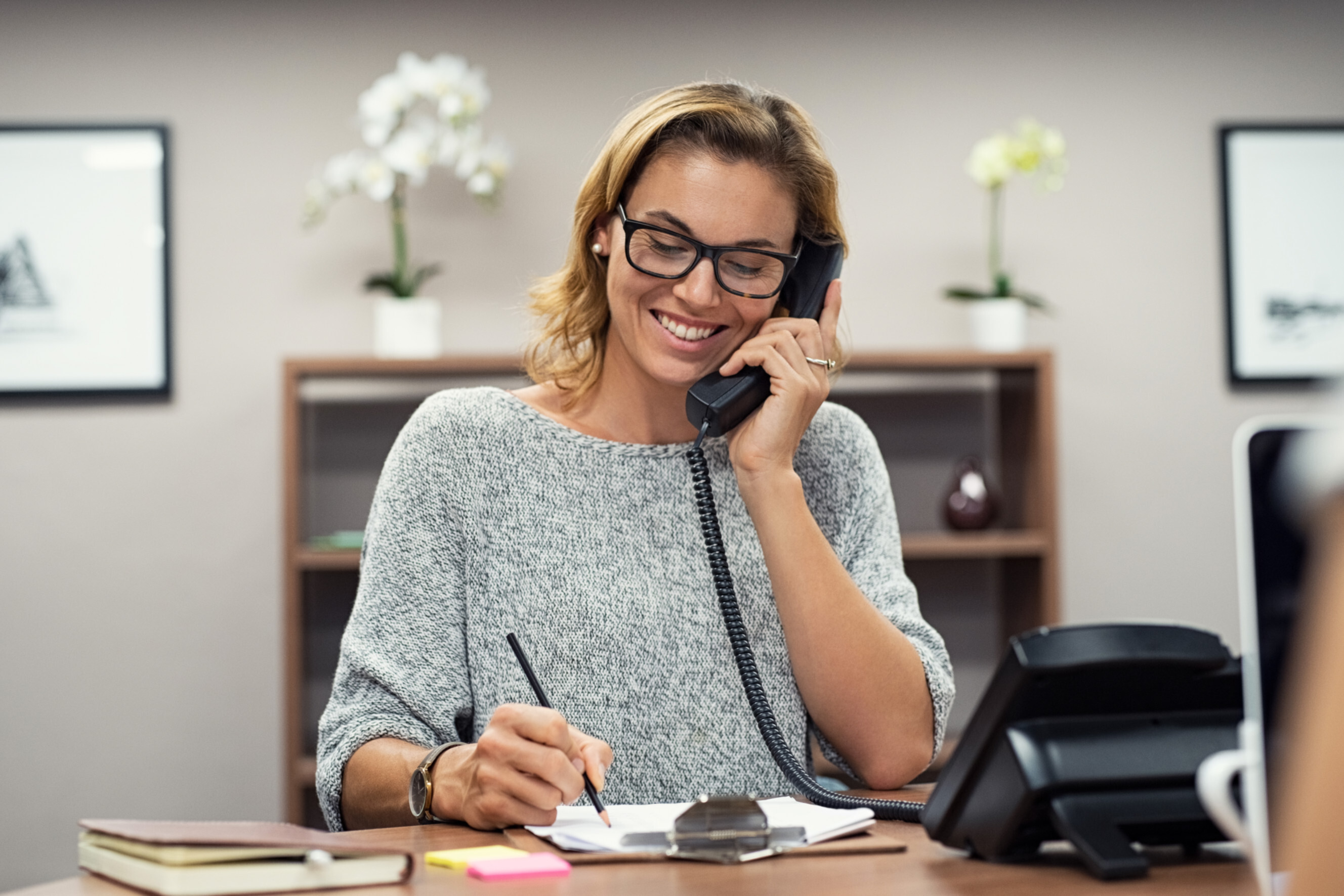 Friendly personal assistant talking on the phone and taking notes at a desk, showcasing professional personal assistants near you.