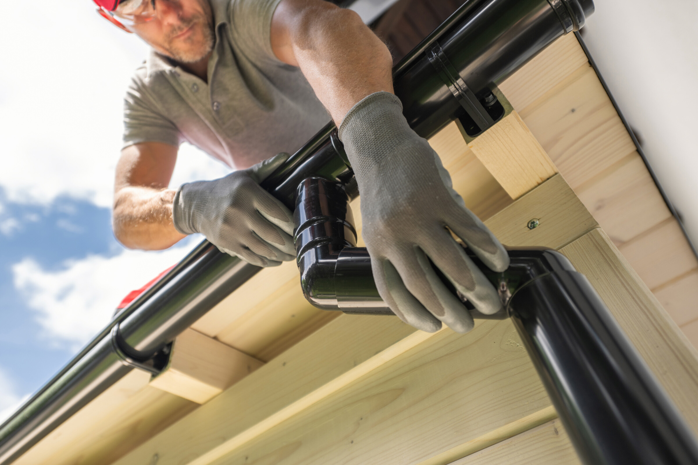 A worker wearing gloves and safety glasses installs a black gutter system on a residential roof. 