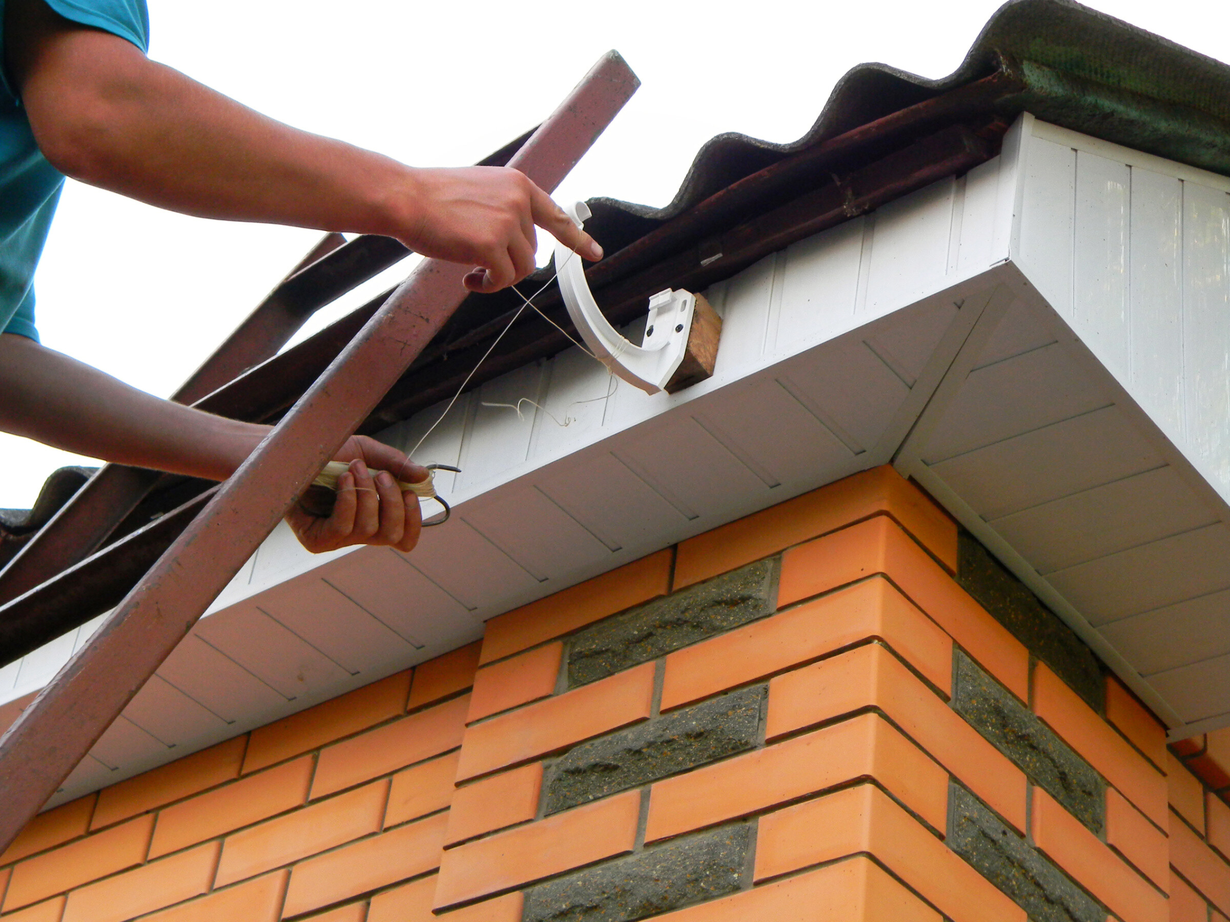 A building contractor is installing rain gutter system, fitting brackets on the top of fascia board, setting up a level line string for the correct positioning of the rain gutter.