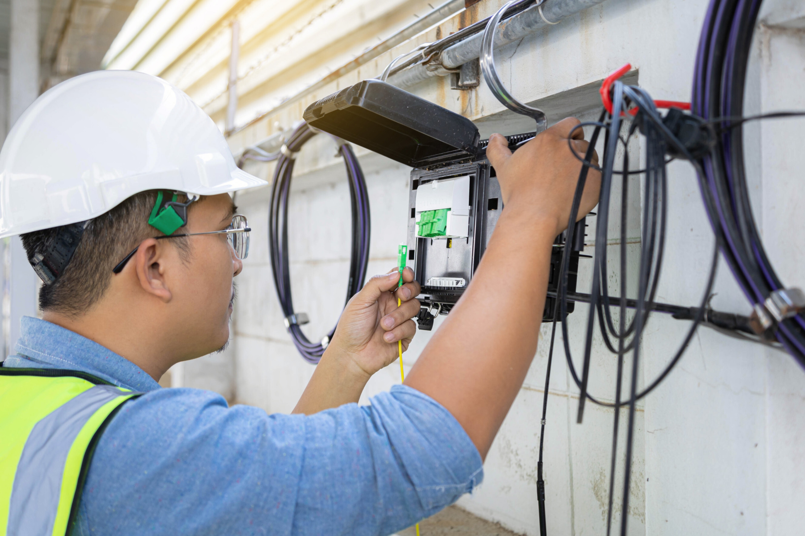 Qualified NBN electrician wearing a safety helmet and high-visibility vest, installing or repairing an NBN connection box with fiber optic cables, representing expert NBN electrician services near you.