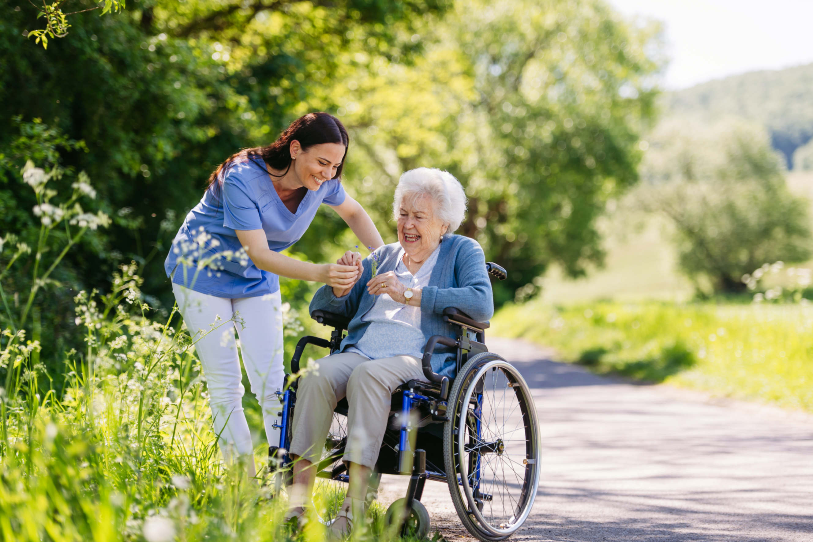 Caregiver assisting an elderly woman in a wheelchair during a nature walk.