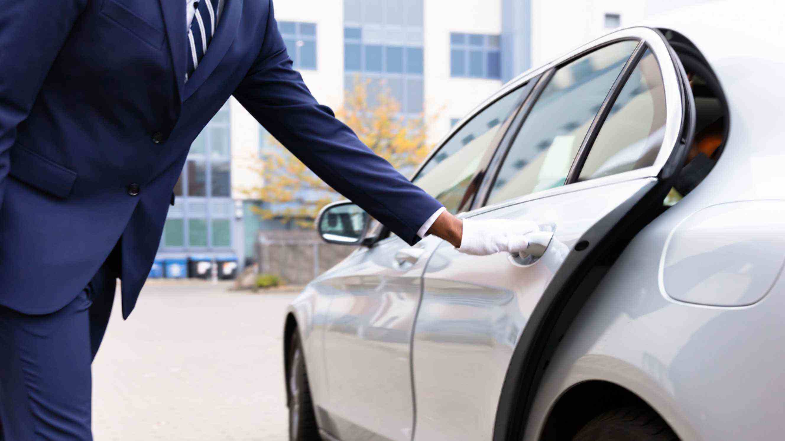 A professional valet in a navy suit and white gloves opening the door of a sleek silver car, representing premium car valet services near you.