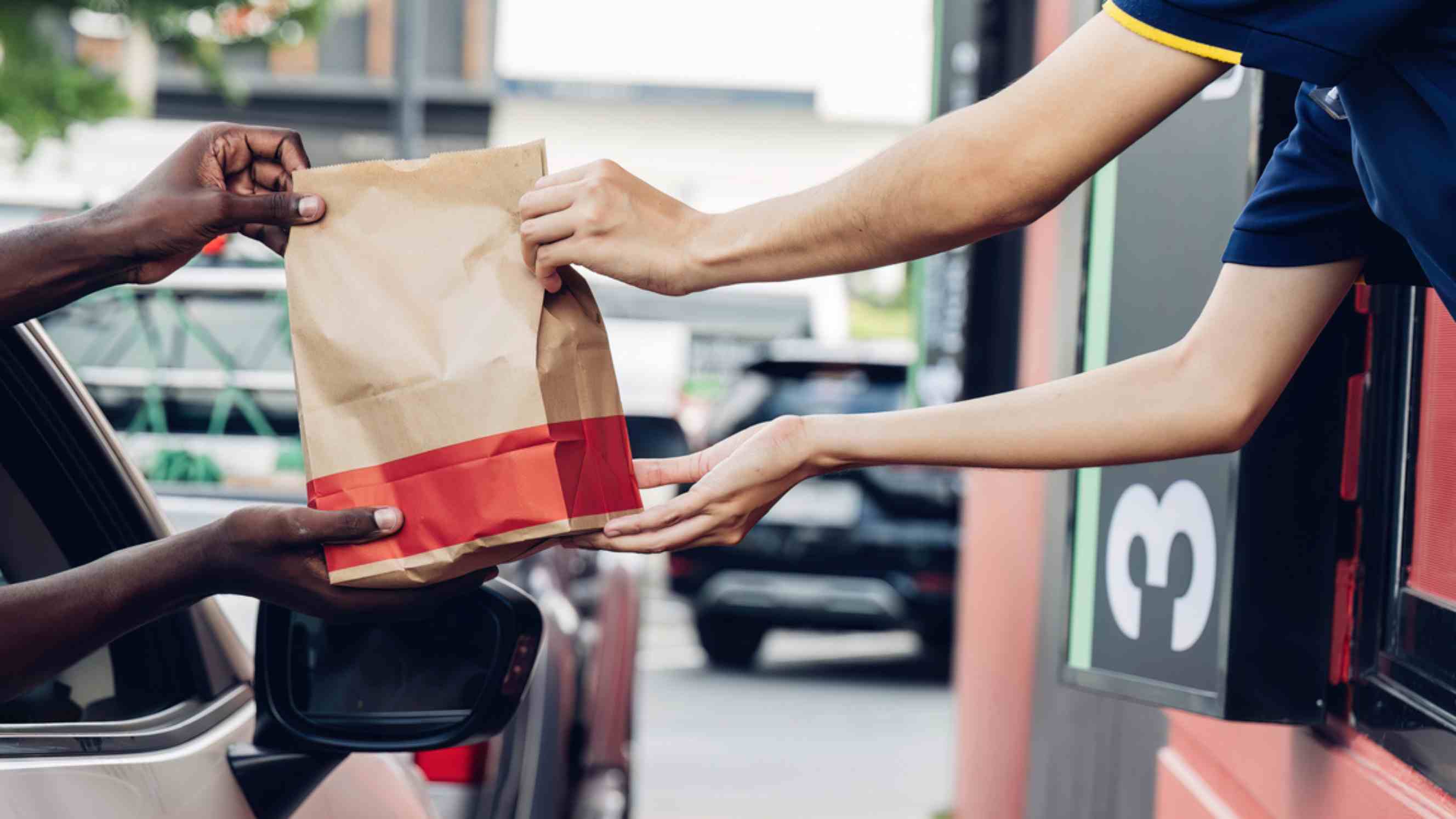 A fast food employee handing a paper bag of food to a customer in a car at a drive-thru window, representing convenient fast food delivery services near you.