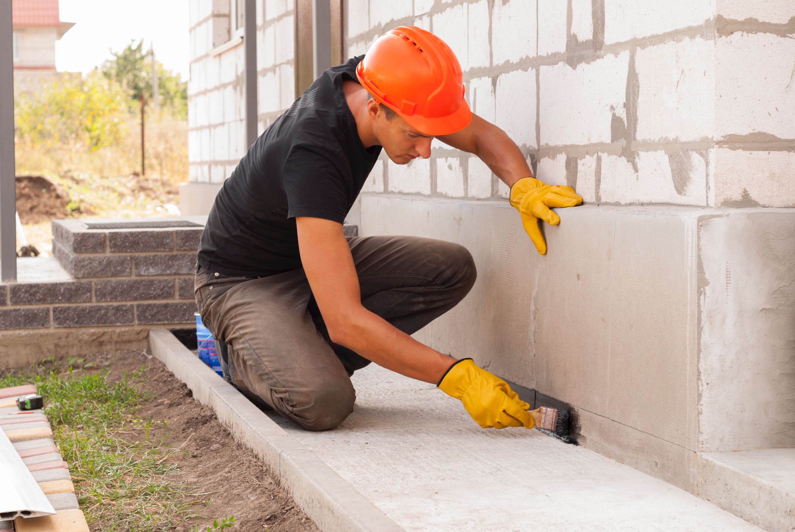 Worker applies bitumen mastic on the foundation representing basement leak repair.