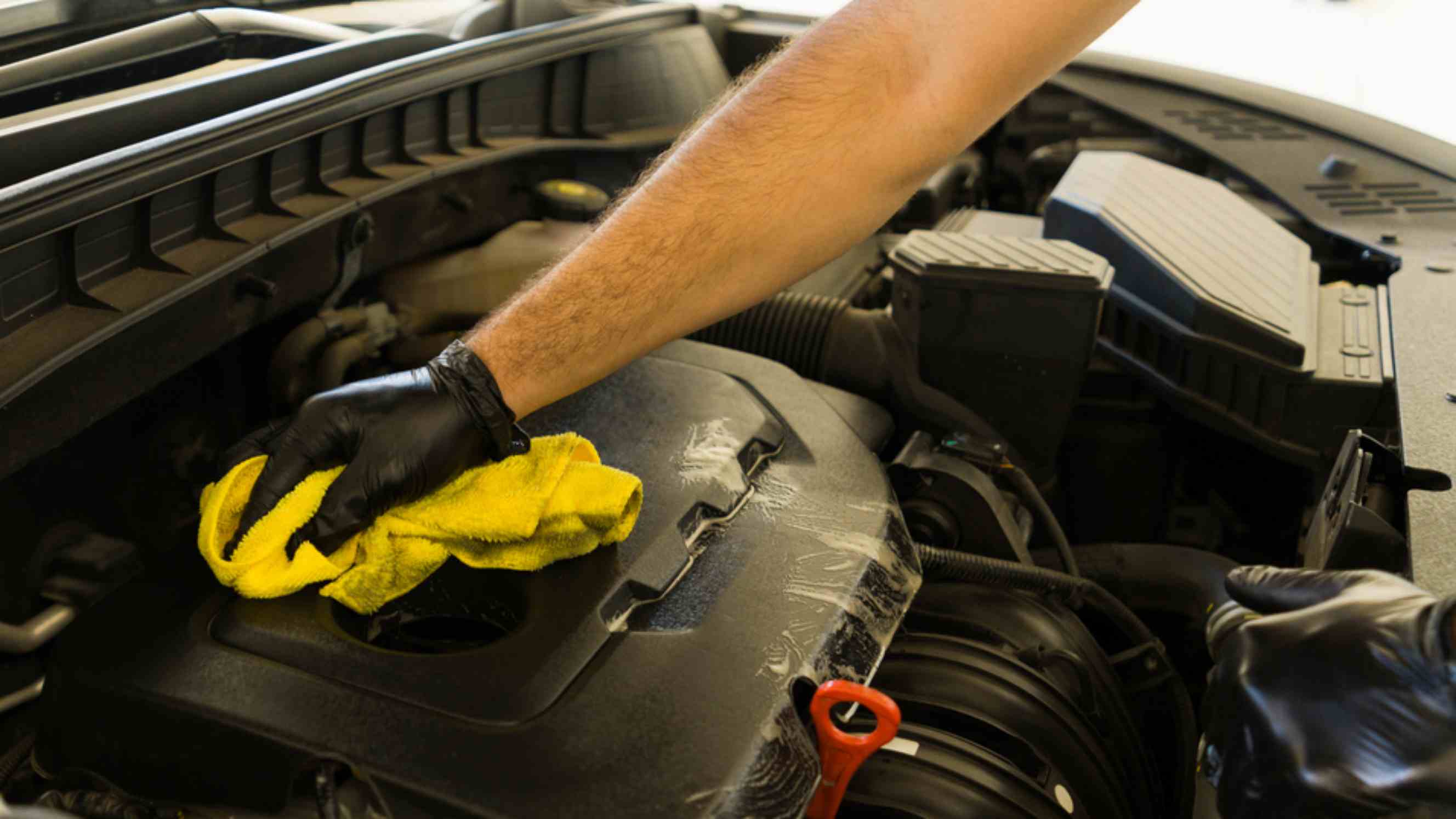 A technician performing car engine steam cleaning near you, using a yellow microfiber cloth to wipe down the engine cover. 