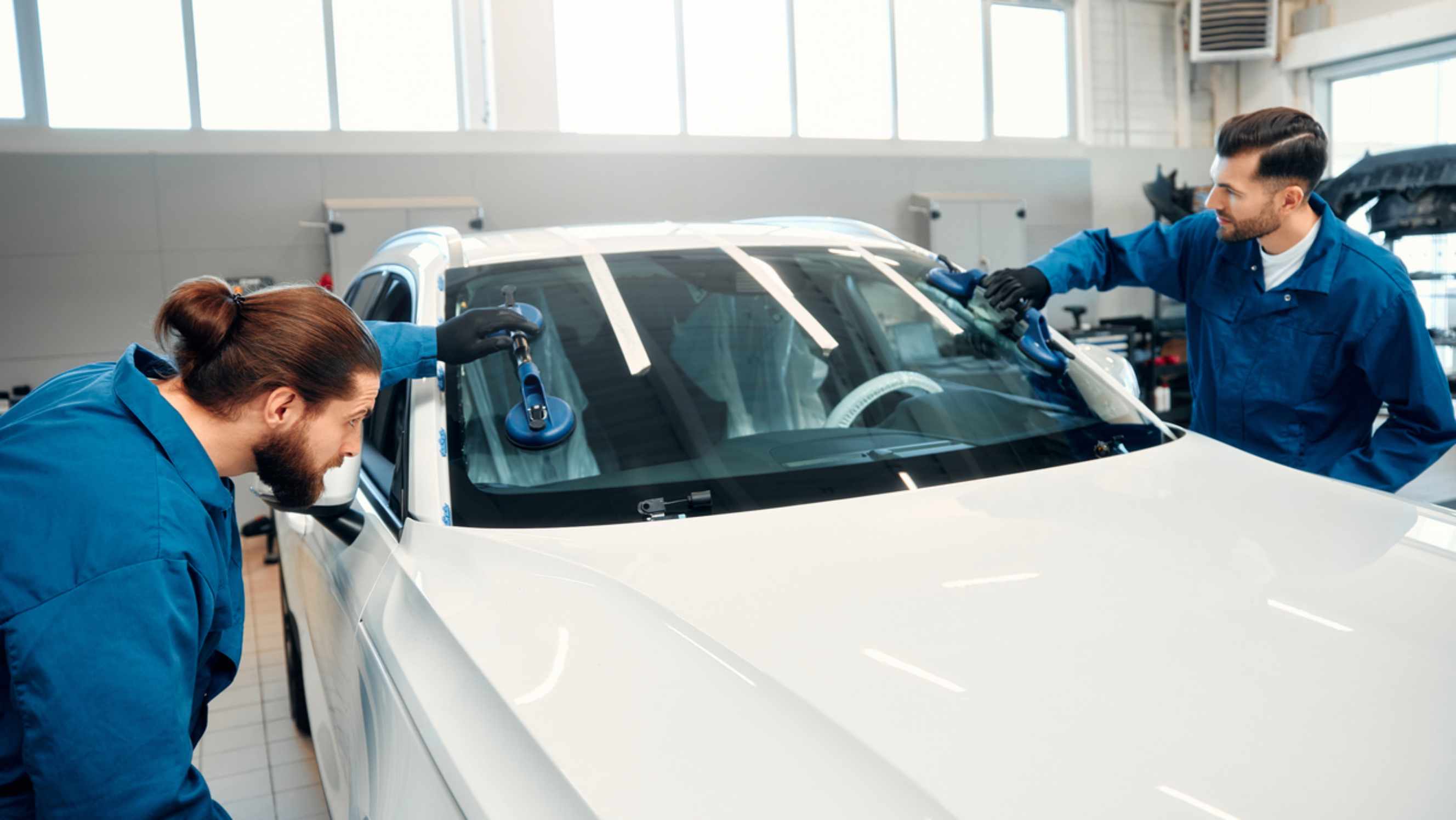 Two technicians performing car glass repair near you, using suction cups to carefully handle and replace the windshield of a white vehicle. 