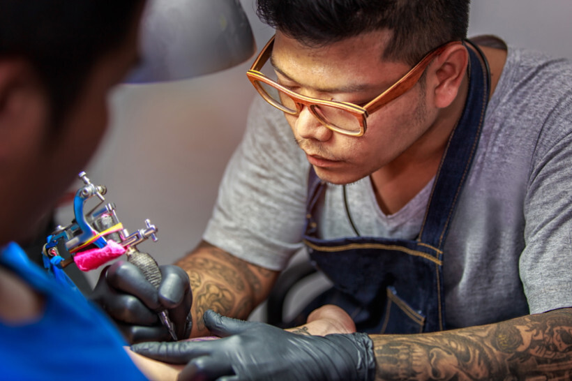 how much do tattoos cost - Professional tattoo artist with glasses inking a detailed forearm tattoo in a vibrant tattoo studio