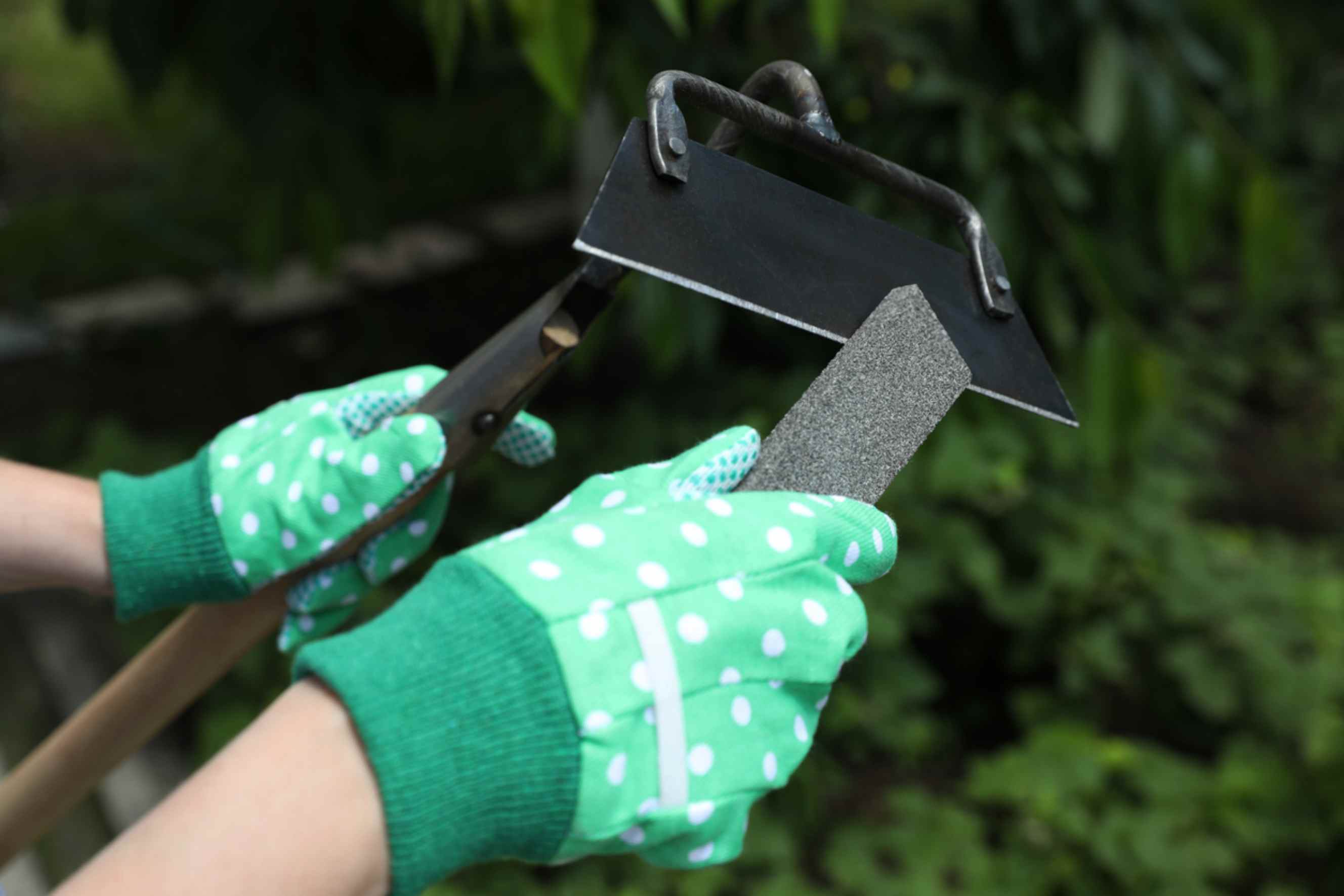 A person wearing green gardening gloves sharpening a hoe, representing garden tool sharpening near you