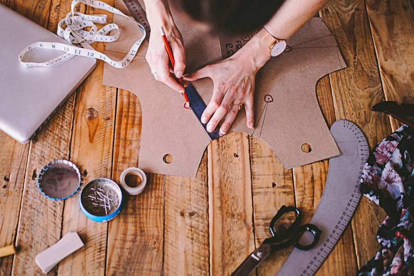 how to make money sewing - Hands working on a garment pattern surrounded by tailoring tools and fabric