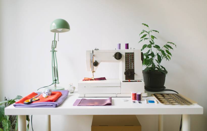 how to make money sewing - Sewing machine on a white desk with fabric, threads, and a green desk lamp
