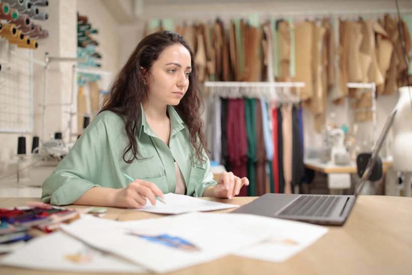 how to make money sewing - Woman working on fashion design sketches at a desk with a laptop in a sewing studio
