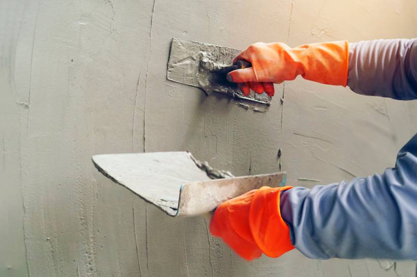 A person wearing orange gloves uses a trowel to apply fresh concrete plaster onto a wall. - lime mortar vs cement mortar