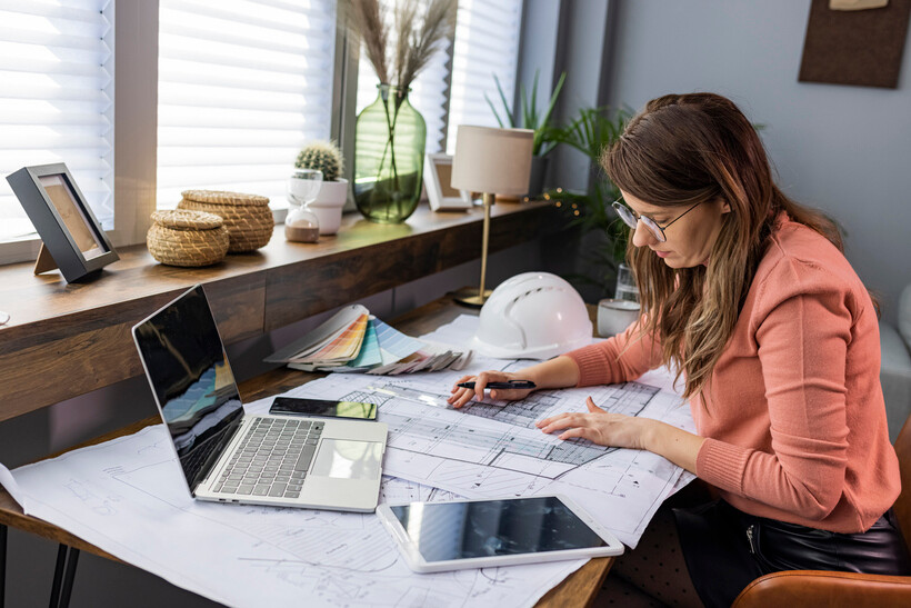 Female architect working on blueprints at a desk with a laptop and tablet. - architect vs engineer