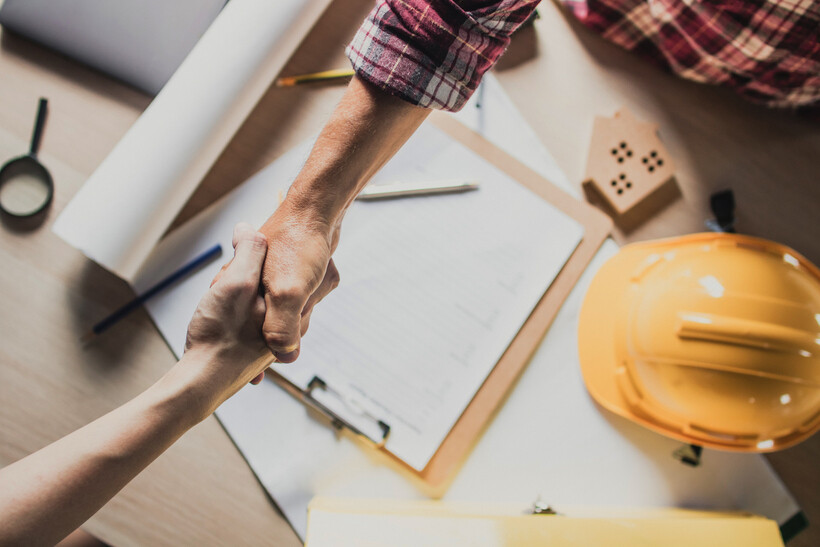 Close-up of a handshake over construction plans and a hard hat on a desk. - renovate vs refurbish