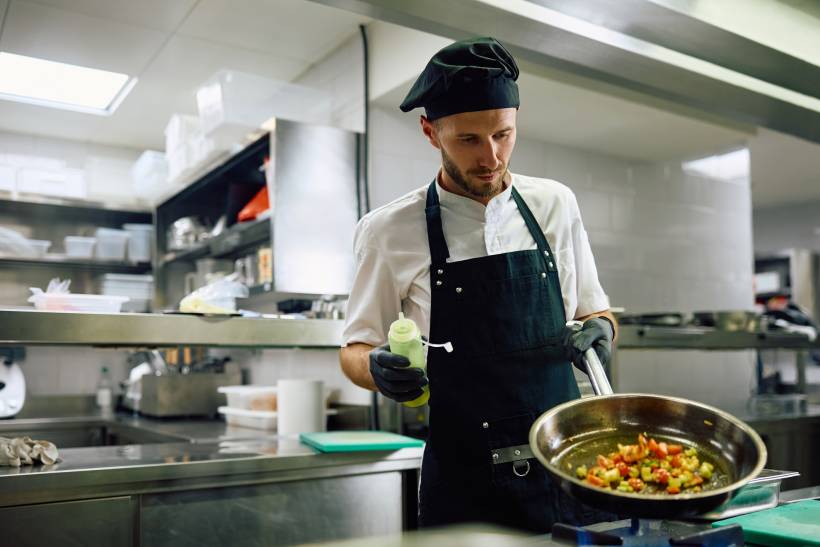 A cook in a commercial kitchen holds a frying pan over a stove while adding oil with a squeeze bottle. - cook vs chef