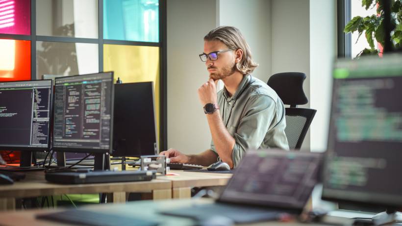 A male programmer is focused on his work, surrounded by computer monitors displaying code. - software developer vs web developer