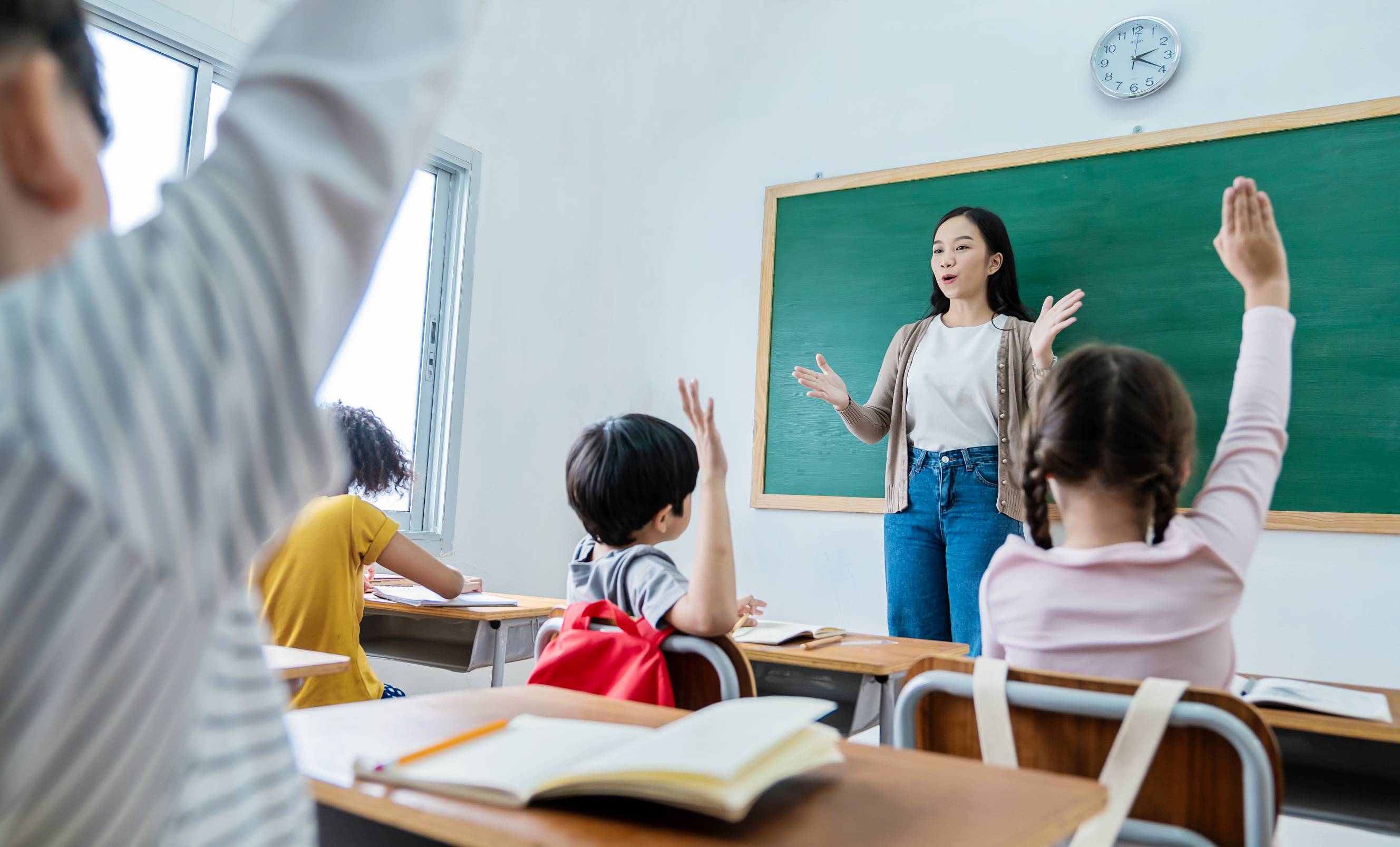 A teacher interacts with her students in a classroom, where several children have raised their hands to answer a question.