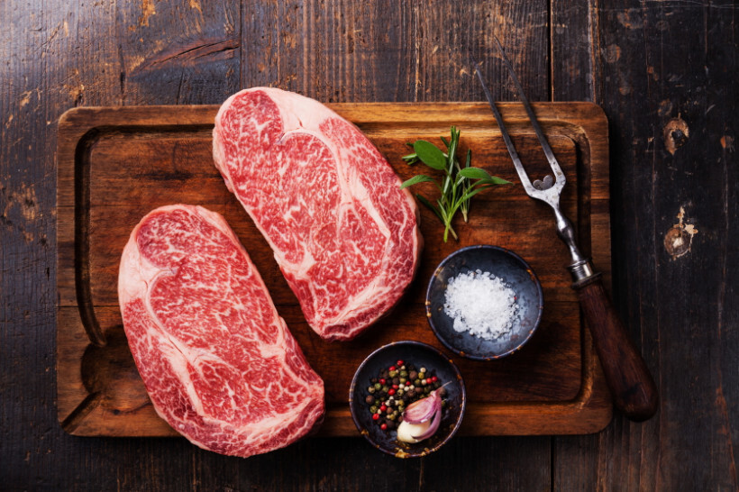 scotch fillet vs porterhouse - Two raw scotch fillet steaks with heavy marbling, salt, and peppercorns on a rustic wooden board.