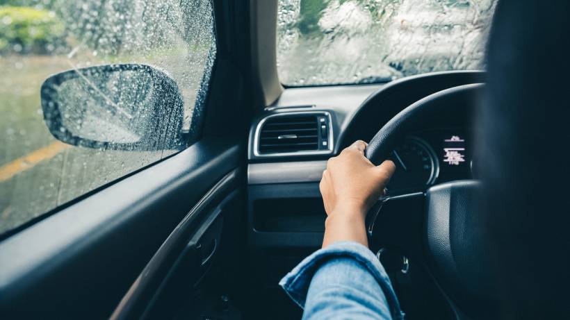 A driver's hands grip the steering wheel as rain streams down the car window on a gloomy day. - make money with your car