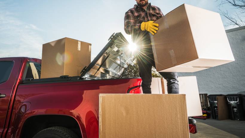 A person wearing work gloves loads a large cardboard box into a red pickup truck. - how to make money with your car