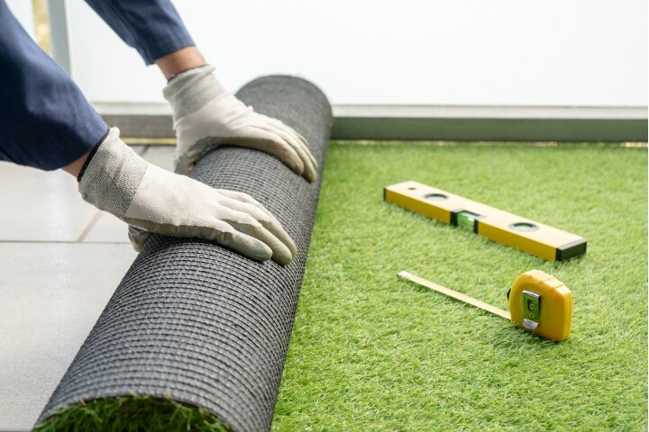 Person wearing gloves unrolling artificial grass on a patio, with a tape measure and level tool nearby.
