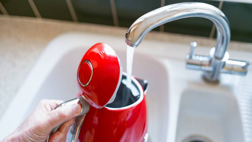 Hand rinsing bright red electric kettle under a chrome kitchen faucet - descale a kettle