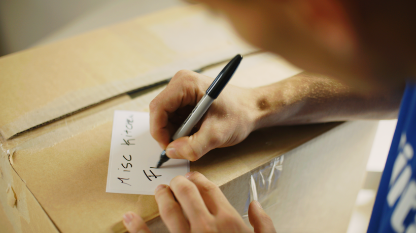 Close-up of a hand writing "Misc Kitchen" on a label attached to a cardboard box. - how to declutter when moving