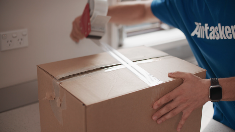 Man wearing a blue shirt taping a cardboard moving box with clear packing tape. - how to declutter when moving