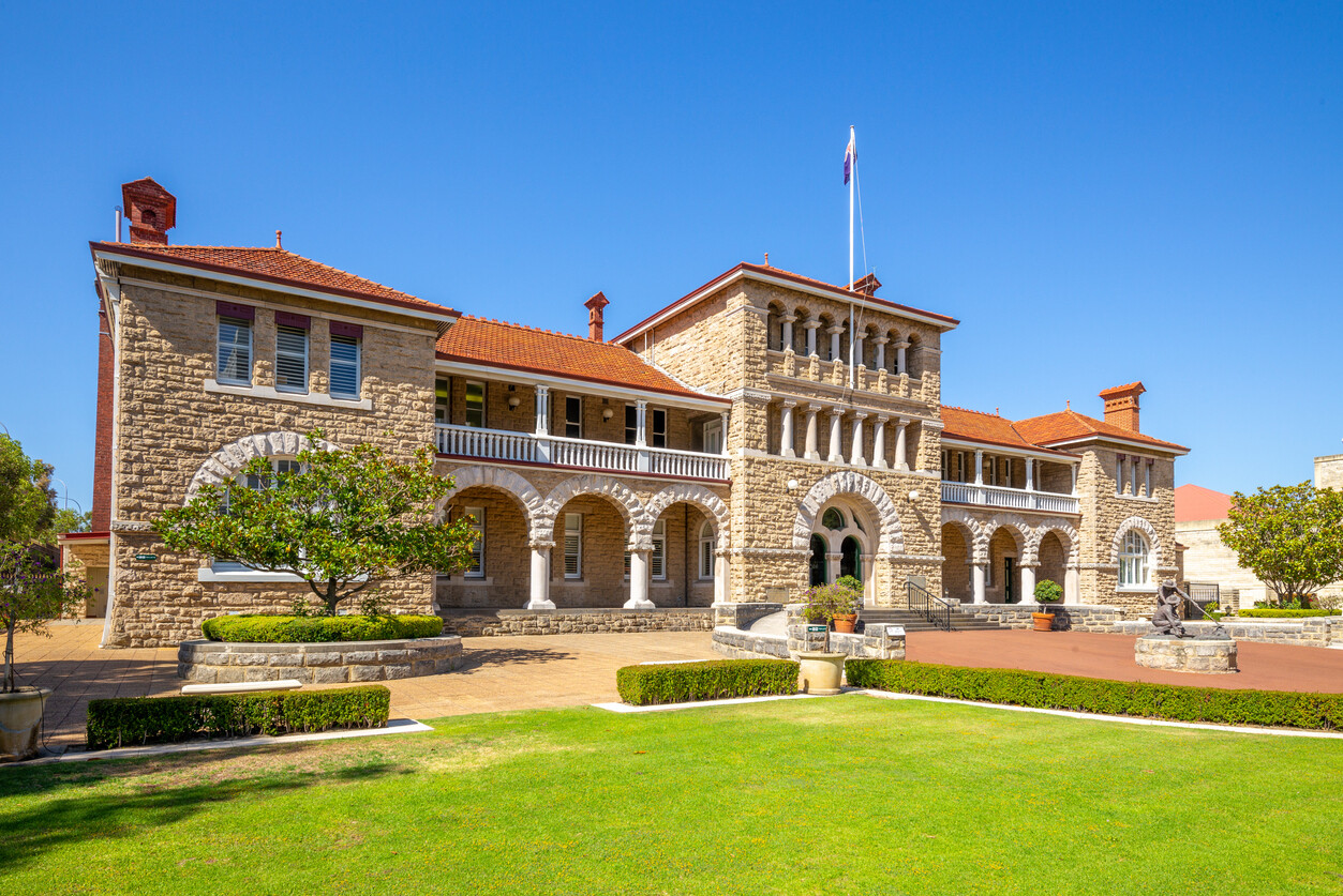 The historic Perth Mint building in Western Australia, showcasing its sandstone exterior, arched verandas, red-tiled roof and landscaped grounds under a clear blue sky