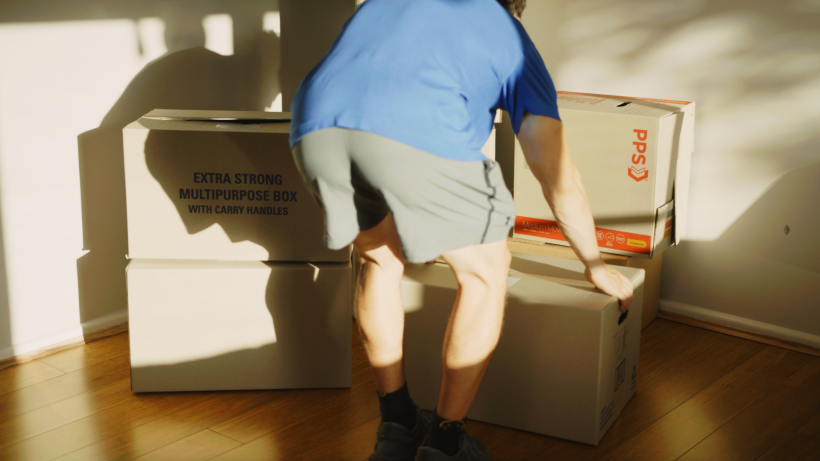 Man lifting a large cardboard moving box off a wood floor in a sunny room - how to pack fragile items