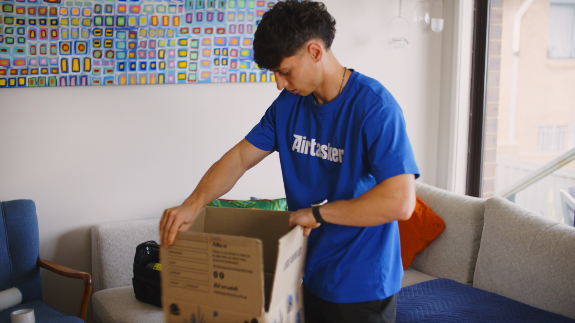 A man in a blue Airtasker shirt assembling a cardboard moving box in a living room.